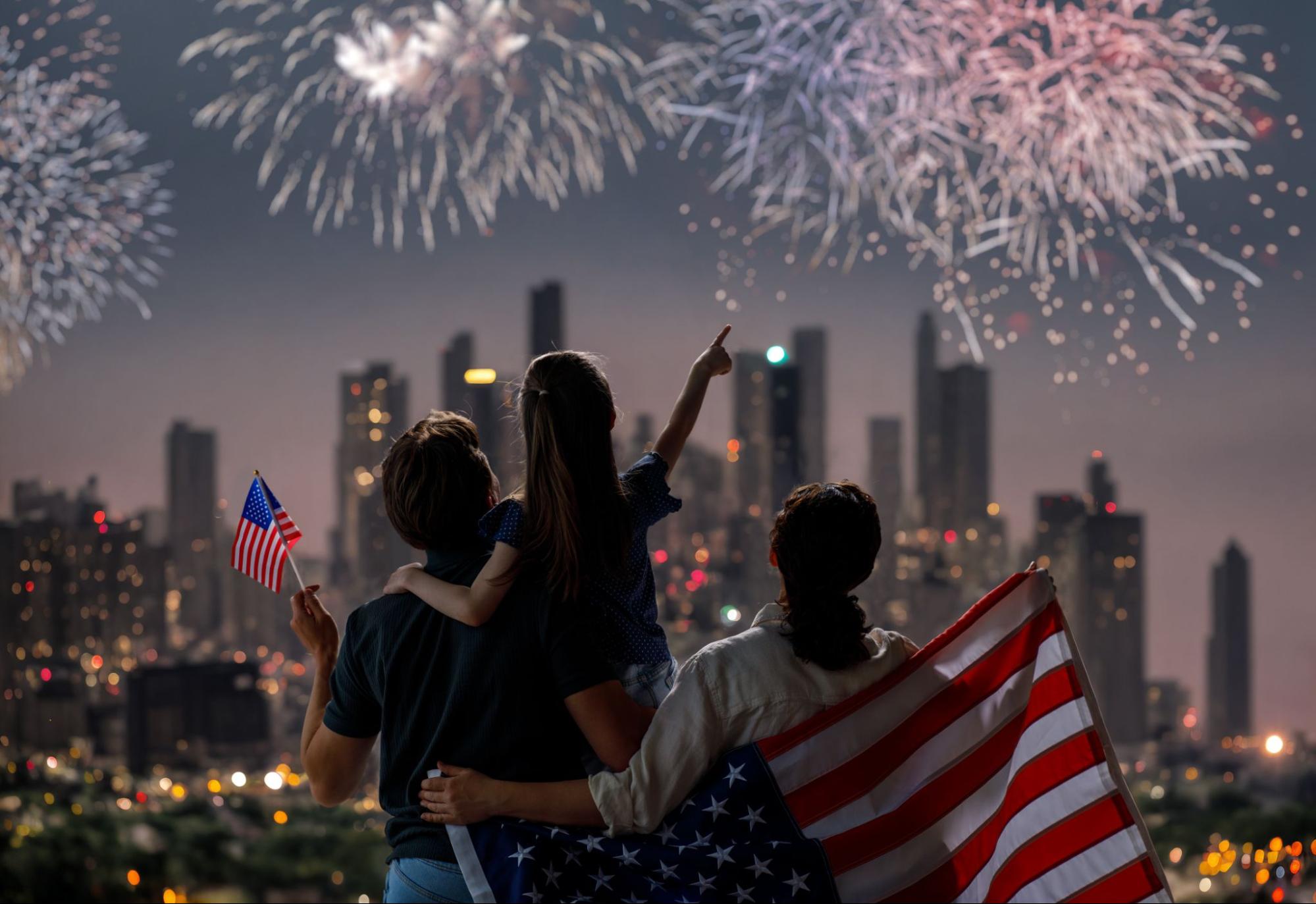 A family watching a fireworks display on Independence Day while holding American flags.