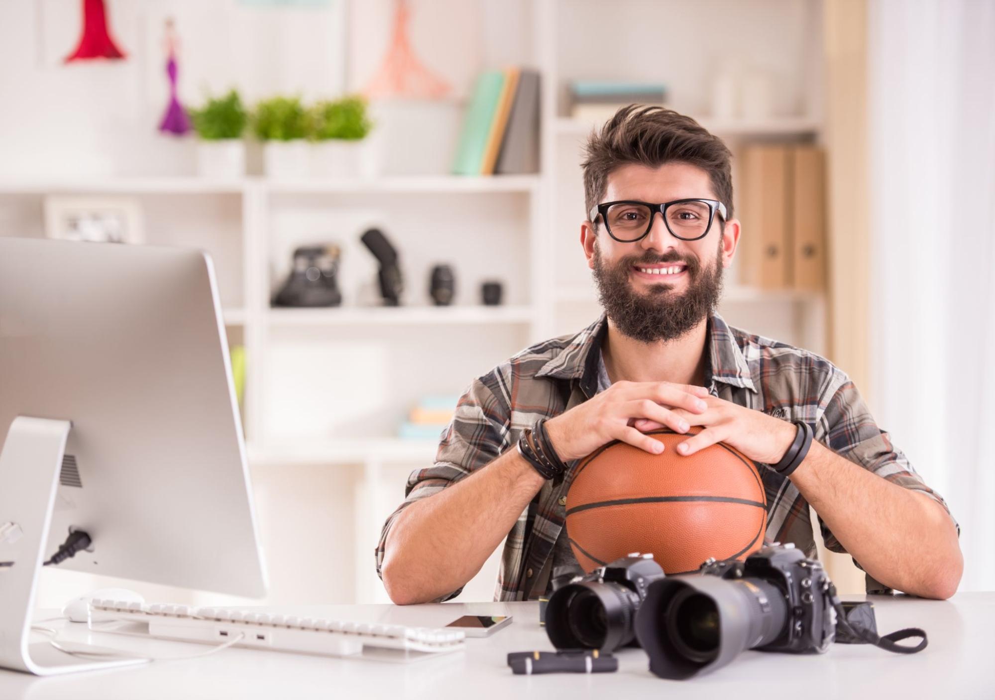 Man smiling next to a computer, basketball, and camera. 