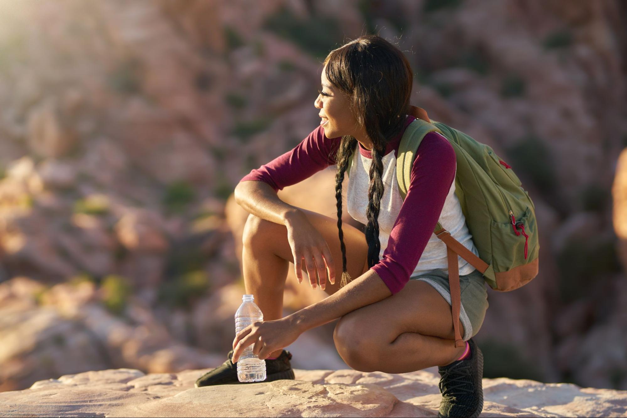 A woman with braids is wearing a baseball shirt and a backpack, crouching atop a cliff.