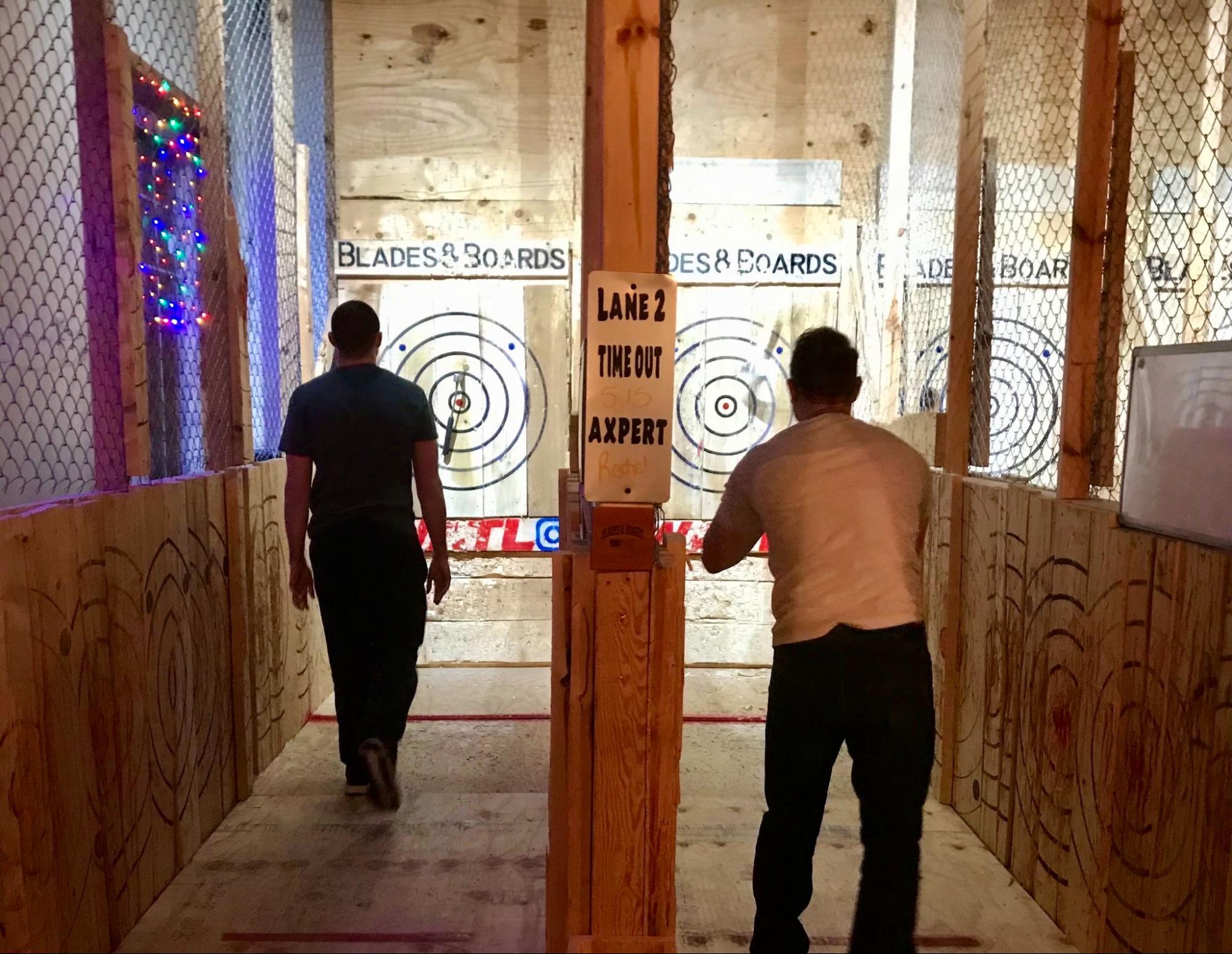 Young men actively throwing axes at targets in an indoor facility, focused and determined, with wooden walls and safety barriers in the background.