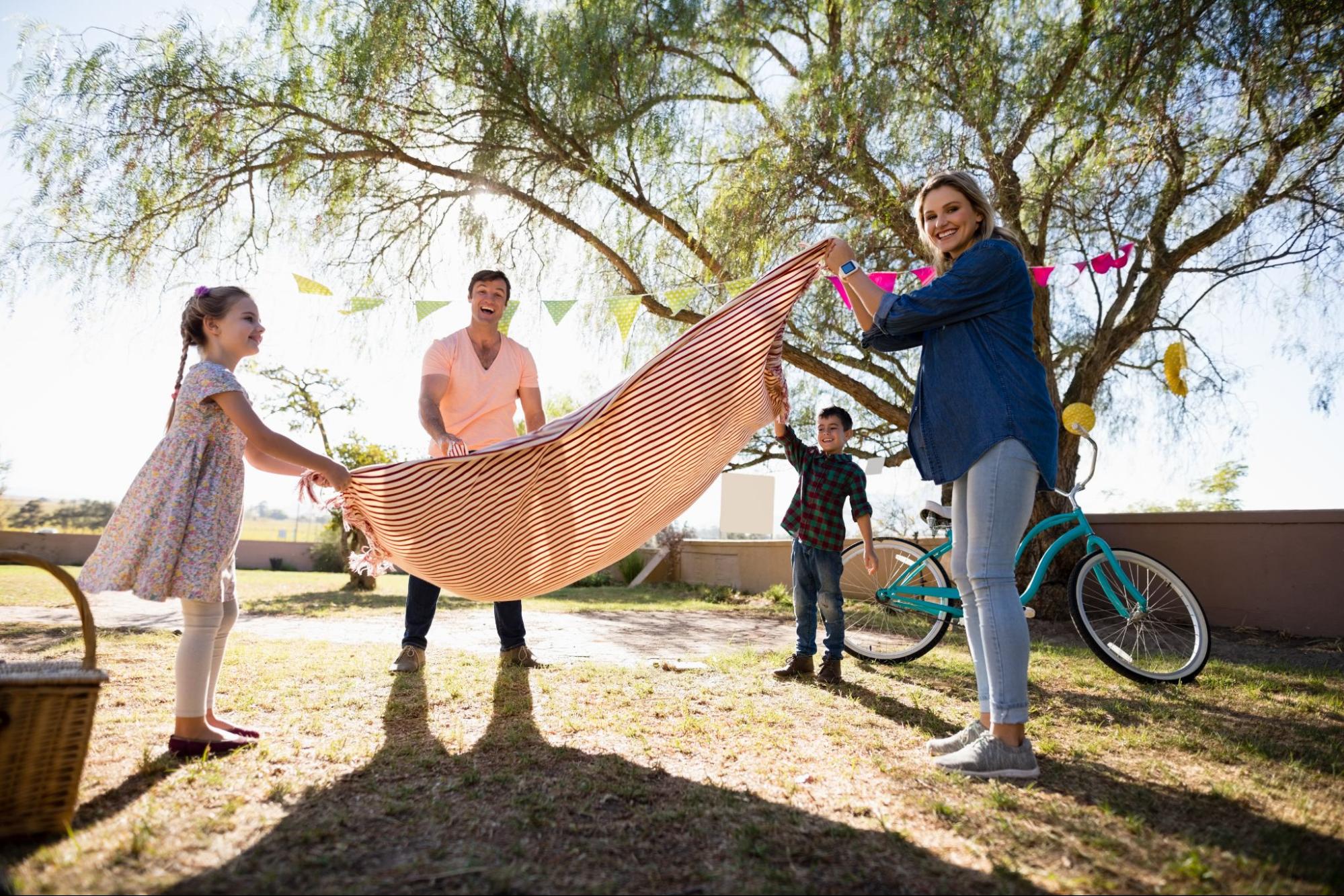 Two adults and two children unfolding a blanket next to a tree and bike. 