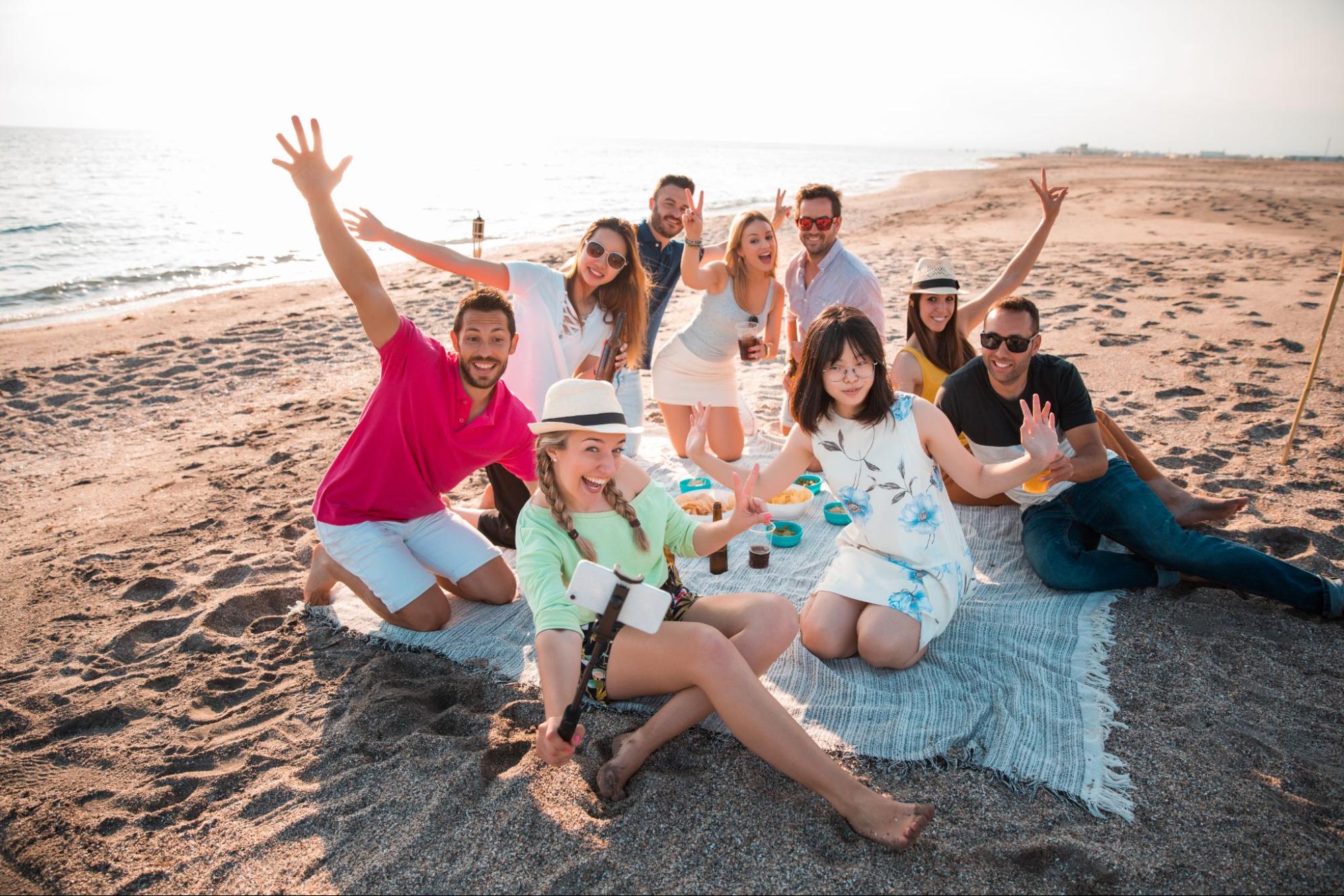 A group of friends posing happily on a beach mat spread on the sand, with some seated and others kneeling, all smiling and enjoying a relaxed, joyful moment together.