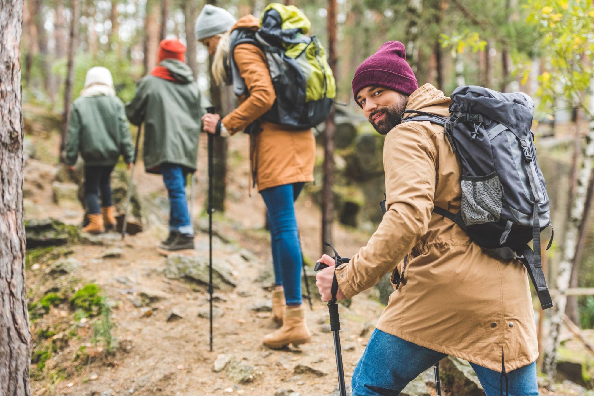 A smiling man in sharp focus, looking at the camera while trekking with his family in an autumn forest.