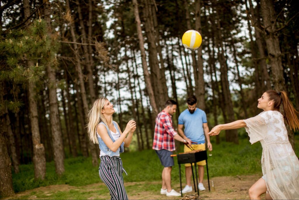 Two girls playing volleyball next to two men grilling.