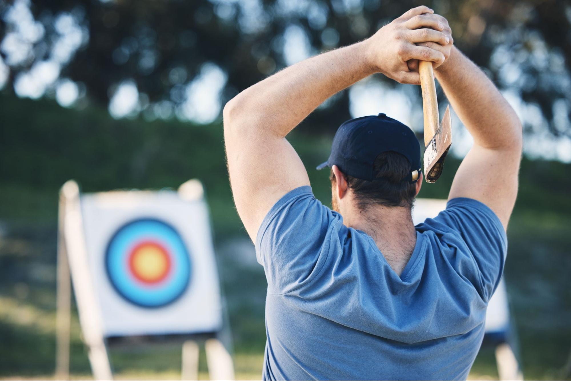 A man at the start of an axe-throwing game prepares to throw toward a circular target board at a sports range.